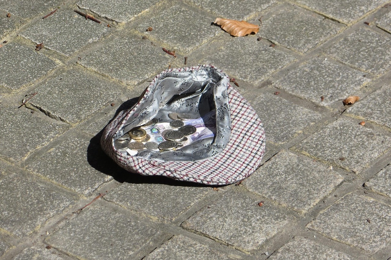 A flat cap on a cobbled floor containing cash.